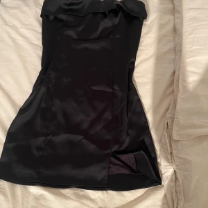 Svart axelbandslös kort klänning från Gina Tricot  - Snygg svart axelbandslös klänning i satinliknande material med liten slits framtill. Perfekt för dig som vill ha en stilren och elegant look. Klänningen har en figurnära passform och en enkel men trendig design.