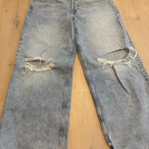 Ljusblå slitna jeans med hål - Säljer ett par ljusblå jeans med rejält slitna hål över båda knäna. Jeansen har en rak passform och klassisk femficksdesign. Perfekta för dig som gillar en avslappnad och trendig streetstil.