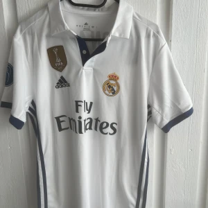 Real Madrid Ronaldo 7 matchtröja - Snygg vit Real Madrid fotbollströja från Adidas med Ronaldo 7-tryck på ryggen. Tröjan har klubbmärke, FIFA-märke och Champions League-patchar på ärmarna. Klassisk krage med knappar och mörkblå detaljer. Materialet är lätt och andas, perfekt för fotboll eller supporterliv.