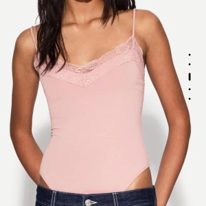 Rosa bodysuit med spetsdetaljer - Supersöt ljusrosa bodysuit med tunna axelband och v-ringning dekorerad med spets. Toppens material är mjukt och stretchigt, perfekt för lager-på-lager eller som statement-piece. Passformen är tight och modellen är ärmlös för en clean och feminin look. ENDAST ANVÄND EN GÅNG säljer pga köpt i fel storlek🩷