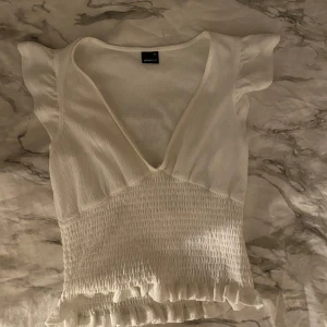 Vit v-ringad topp  - Supersöt vit topp från Gina Tricot i storlek XS. Den har v-ringning, korta volangärmar och smockad midja med volangkant nertill. Jag tycker inte att den passar mig så bra så därför säljer jag den! 🤍den är använd fåtal gånger och är i superbra skick! 💋