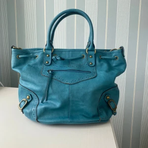 Turkos axelväska i skinn med detaljer - Snygg turkos blå axelväska med metalldetaljer och dekorativa sömmar. Väskan har både handtag och justerbar axelrem som kan stoppas in. Perfekt storlek, ca 41 cm bred och 25cm hög så dator m.m får lätt plats! Skicket är väldigt bra bara små defekter så som lätta missfärgningar inuti (se bild) och något litet skav på utsidan☺️