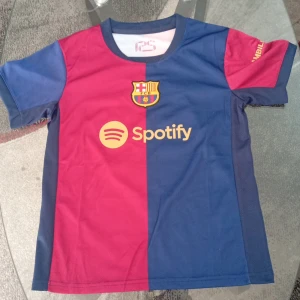 FC Barcelona Lamine Yamal matchtröja set - FC Barcelona matchtröja och shorts med Lamine Yamal 19-tryck. Tröjan är delad i rött och blått med klubbmärke och Spotify-logga framtill, UNHCR-logga och namn/nummer bak. Korta ärmar, sportigt material och klassisk passform. Shorts i blått med klubbmärke och nummer.
