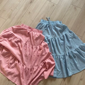Pastellfärgade volangkjolar med knyt - Två superfina volangkjolar i pastellrosa och ljusblått. Kjolarna har resår i midjan med knytband och är sydda i ett luftigt bomullstyg. Perfekta för varma dagar och ger en söt och avslappnad vibe. 50kr för båda.