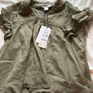 Olivgrön blus med volangärm från Gina tricot - Supersöt olivgrön blus från Gina tricot med korta volangärmar och fina spetsdetaljer. Blusen har rynkade partier upptill och en avslappnad passform. Perfekt för dig som gillar en boho-vibe och mjuka material. Aldrig använt den förut och lappen är fortfarande kvar.