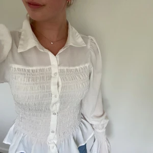 Vit blus med smock och volang - Supersöt vit blus med smockad midja och volangkant nertill. Långa ärmar med puff och smock vid ärmslut, klassisk krage och knappar framtill. Tunt, lätt tyg som känns luftigt och fräscht. Perfekt till jeans eller kjol för en trendig look.