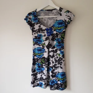 Blommig kortklänning från Lady's Rose - Supersöt kortklänning med blommigt mönster i blått, svart och grönt på vit botten. Klänningen har rund halsringning, korta ärmar och dekorativa blå knappar framtill. Perfekt för dig som gillar färg och mönster!  Helt skick dock obehandlade fläckar & noppror