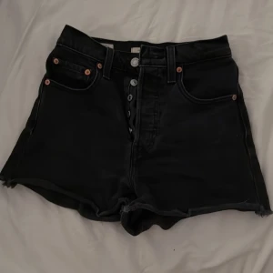 Svarta Levi's Ribcage shorts jeans - Svarta högmidjade jeansshorts från Levi's modell Ribcage. Klassisk femficksdesign, råa kanter nertill och knappgylf. Snyggt Levi's-märke bak i läder och röd logga på bakfickan. Perfekta för en cool och avslappnad stil.
