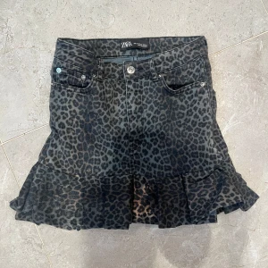 Leopardmönstrad jeanskjol från Zara - Cool kort jeanskjol från Zara med leopardmönster i mörka toner. Kjolen har volang nertill, klassiska fickor fram och bak samt knapp och dragkedja framtill. Perfekt för dig som vill sticka ut med en edgy vibe.