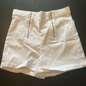 Vita shorts med dragkedjor FB Sister - Snygga vita shorts från FB Sister i storlek S. De har hög midja och två coola silverfärgade dragkedjor framtill med ringdetaljer. Materialet känns som bomull och shortsen har en clean, stilren look med uppvikta benslut. Perfekta för sommardagar!