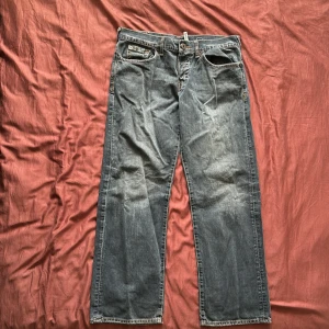 Guess Jeans blå raka jeans W36 L30 - Klassiska blå jeans från Guess Jeans med raka ben och snygga kontrastsömmar. Jeansen har fem fickor, läderpatch bak och är tillverkade i 100% bomull. Perfekta för dig som gillar en avslappnad och tidlös look.
