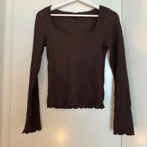 Brun långärmad topp från Gina Tricot - Mörkbrun långärmad topp från Gina Tricot i storlek S. Tröjan har en fyrkantig urringning och är tillverkad i ett stretchigt, ribbat material med vågiga kanter vid ärmslut och nederkant. Perfekt att styla med jeans eller kjol för en trendig look.