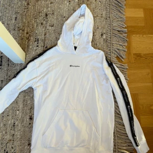 Vit Champion hoodie med logoband - Snygg vit hoodie från Champion med svart logoband längs ärmarna och broderad logga på bröstet. Klassisk känguruficka framtill och huva. Perfekt för en chill och sportig look.