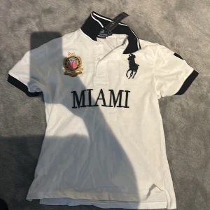 Vit Ralph Lauren Miami t-shirt - Vit t-shirt från Ralph Lauren med svart krage och ärmslut, stor broderad logga och texten MIAMI framtill. Snygg detalj med klubbmärke på bröstet och stor siffra 5 på ryggen. Klassisk pikélook med korta ärmar och sportig vibe.
