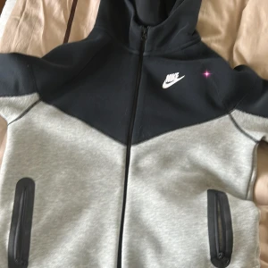 Nike hoodiejacka svart/grå  - Nike hoodiejacka i svart och ljusgrått med dragkedja framtill och två fickor med svarta detaljer. Jackan har en sportig look, huva och klassisk Nike-logga på bröstet. Perfekt för chill eller när du är på språng.