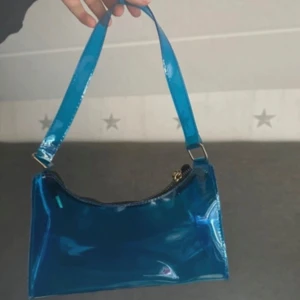 Blå axelväska i glansigt material - Säljer en cool blå axelväska med glansig finish och enkel design. Väskan har ett brett axelband och stängs med dragkedja upptill. Perfekt för att addera färg till din outfit.