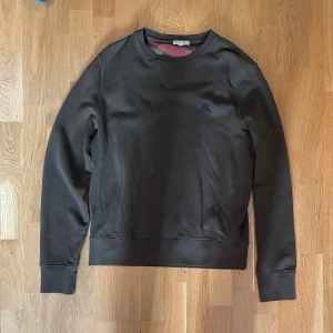 Mörkgrå sweatshirt från Burberry - Snygg mörkgrå sweatshirt från Burberry med broderad logga på bröstet och klassiskt rutigt tyg på insidan av kragen. Tröjan har rund halsringning och långa ärmar, perfekt för dig som gillar stilrena och exklusiva plagg.