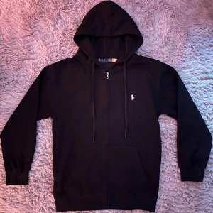 Svart zip hoodie från Polo Ralph Lauren - Svart hoodie från Polo Ralph Lauren med klassisk vit broderad logga på bröstet. Tröjan har huva med snörning, dragkedja framtill och känguruficka. Tillverkad i mjuk bomull, perfekt för chill dagar. Snygg och enkel att matcha med allt. Kvitto kan fås med om man vill