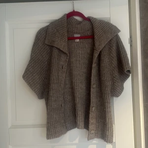 Beige stickad kofta med kort ärm - Mysig beige stickad kofta med korta ärmar och bred krage. Koftan har grov ribbstickning och knappar framtill, perfekt för lager-på-lager. Avslappnad passform och mjukt material som känns skönt mot huden.