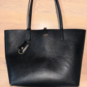 Svart shoppingväska från Lauren Ralph Lauren - Stilren svart shoppingväska i präglat skinn från Lauren Ralph Lauren. Väskan har två långa handtag, guldfärgade detaljer och medföljande grå innerficka med RL-logga och dragkedja. Perfekt för dig som vill ha plats för allt och ändå se classy ut.