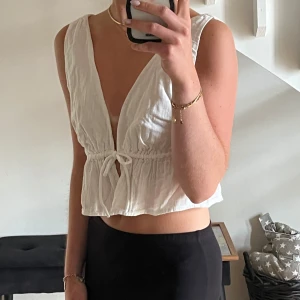 Vit croppad linnetopp med knyt - Säljer en vit croppad linnetopp med djup v-ringning fram och bak. Toppen har knyt i midjan och volangkant nedtill och är oanvänd. Perfekt till stranden eller en mysig sommarkväll.