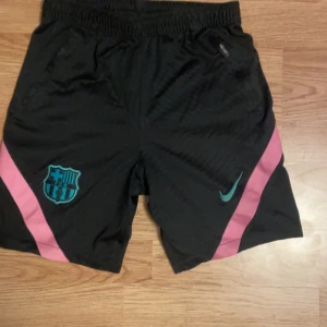 FC Barcelona x Nike svarta shorts - Svarta FC Barcelona shorts från Nike med rosa diagonala ränder på sidorna och turkosa detaljer. Broderad FCB-logga och Nike Swoosh framtill. Elastisk midja och dragkedjeficka. Perfekta för hemma eller träning.