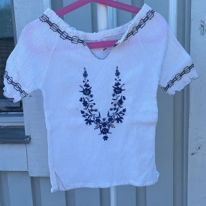 Vit blus med broderi och offshoulder - Superfin vit blus i bomull med offshoulder-modell och korta ärmar. Blusen har mörkblå broderade detaljer längs halsringning och ärmslut samt ett snyggt blommigt broderi framtill. Tyget är lätt skrynklat och känns luftigt, perfekt för varma dagar.