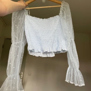 Ljusblå drömmig topp - Supersöt ljusblå topp med smockad kropp och volangkant nertill. Ärmarna är långa, puffiga och gjorda i transparent mesh med glittriga stjärnor och månar. Perfekt för dig som vill sticka ut med en drömmig och trendig look. L men passar M