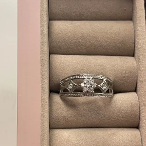 Silverring med glittrande stenar - Snygg silverfärgad ring med glittrande stenar i mitten och på sidorna. Ringen har ett öppet, dekorativt mönster och små detaljer längs kanterna som ger extra bling. Perfekt för dig som gillar unika och eleganta smycken.