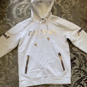 Vit hoodie från Balmain med guldtryck - Exklusiv vit hoodie från Balmain med guldig logga på bröstet. Tröjan har huva med snörning, flera snygga dragkedjor i guld och quiltade detaljer på axlarna. Perfekt statement-plagg för dig som gillar lyxig streetstyle.