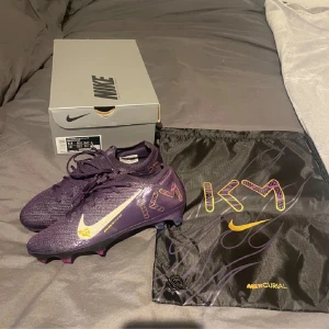 Nike Mercurial Vapor 16 Elite KM FG - Säljer ett par Nike Mercurial Vapor 16 Elite KM FG fotbollsskor i mörklila med vita och gula detaljer. Skorna har snörning, mönstrade KM-loggor och en lätt, syntetisk ovandel. Perfekta för dig som vill sticka ut på planen. Levereras med originalpåse och låda.