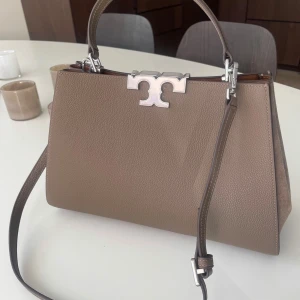 Beige handväska från Tory Burch - Snygg beige handväska från Tory Burch med silverfärgad logga framtill. Väskan har både axelrem och handtag, är tillverkad i skinn med mockadetaljer på sidorna. Stilren och rymlig modell med klassisk form och exklusiv känsla. Endast använd 1 gång. Så helt som ny.