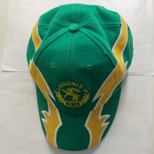 Grön Ljusdals IF keps med blixtar - Snygg grön keps från Ljusdals IF med broderad gul logga framtill och coola gula och vita blixtdetaljer längs sidorna. Justerbar baktill och tillverkad i bomull. Perfekt för dig som vill sticka ut på läktaren eller i vardagen.