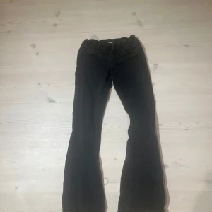 Svarta bootcut jeans med fickdetaljer - Snygga svarta jeans med bootcut passform och klassiska fickor bak med lock och knapp. Jeansen har normal midja med lite stretch. Perfekta för dig som gillar en tidlös look med lite extra edge.