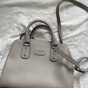 Grå handväska från Michael Kors - Snygg grå handväska från Michael Kors med både handtag och justerbar axelrem. Den är aldrig använd utan har legat i min garderob sen 2017!