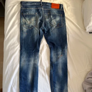 Äkta Dsquared2 blå distressed jeans - Säljer ett par blå jeans från Dsquared2 med coolt distressed och slitna detaljer, färgstänk och hål. Jeansen har klassisk femficksdesign, läderpatch bak och snygg tvättad look. Perfekta för dig som gillar streetstyle och vill sticka ut. (AI)