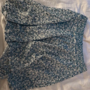 Blommig blå kortkjol med resår - Supersöt blå kortkjol med vitt och svart blommönster. Kjolen har en flowy passform och bred resår i midjan för extra komfort. Perfekt för varma dagar och ger en fräsch vibe till din outfit.