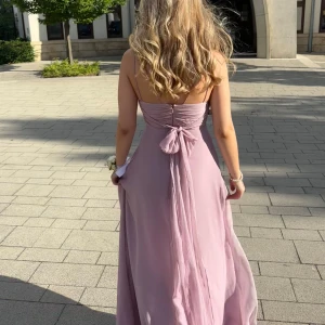 Ljuslila långklänning med rosett - ENDAST ANVÄNT EN GÅNG! Superfin långklänning i ljuslila med smala axelband och v-ringning. Klänningen har en elegant rosett i ryggen och faller snyggt i ett luftigt material. Perfekt för speciella tillfällen.