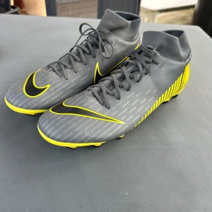 Nike Mercurial grå/gula fotbollsskor - Snygga Nike Mercurial fotbollsskor i grått med gula och svarta detaljer. Skorna har snörning, hög strumpliknande krage och mönstrad sula med dobbar för bra grepp på planen.