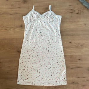 Vit blommig kortklänning med spets - Supersöt vit kortklänning med rosa blommor och tunna axelband. Klänningen har spetsdetaljer vid bysten och längs kanterna, samt en mjuk och glansig känsla i tyget. Perfekt för sommarens varma dagar eller när du vill känna dig extra gullig.