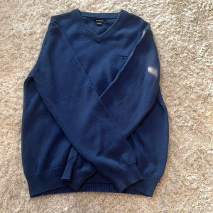  Stickad Blå v-ringad tröja från GANT - Klassisk blå v-ringad tröja från GANT med diskret logga broderad på bröstet. Tröjan är stickad i bomull och har ribbade muddar vid ärmslut och nederkant. Perfekt för dig som gillar en stilren och tidlös look.