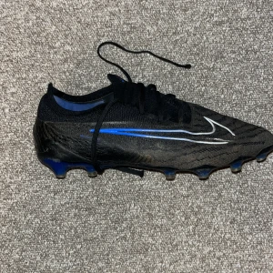 Nike Phantom svart/blå fotbollssko - Nike Phantom fotbollssko i svart med blå detaljer och vit swoosh. Ovandel i syntetmaterial med mönstrad textur för bättre bollkontroll. Plös och snörning för tajt passform. Yttersula med fasta dobbar i blått för bra grepp på planen. Ny pris 3400.