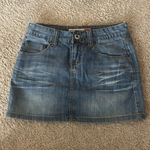 Lågmidjad jeanskjol - Säljer en klassisk blå jeanskjol från D.A.D.E med fem fickor och snygga slitningar framtill. Kjolen har knapp- och dragkedjestängning samt dekorativa bakfickor. Perfekt till sommaren och enkel att matcha med olika toppar.