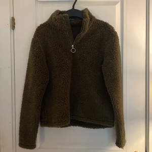 Olivgrön teddytröja med dragkedja från Rut&Circle - Mysig olivgrön teddytröja från Rut&Circle med halv dragkedja och hög krage. Tröjan har lång ärm och är tillverkad i ett fluffigt, fleece-liknande material som håller dig varm under kyliga dagar.