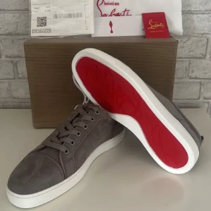 Grå sneakers från Christian Louboutin med röd sula - Säljer ett par gråa sneakers från Christian Louboutin med ikonisk röd sula och vita kanter. Skorna har snörning och rund tå, tillverkade i textil och skinn. Perfekta för dig som vill sticka ut med exklusiva detaljer. (6)