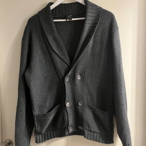 Grå stickad kofta från Jankowich - Mörkgrå stickad kofta från Jankowich med bred sjalkrage och dubbelknäppning framtill. Koftan har två framfickor och ribbade muddar vid ärmslut och nederkant. Perfekt för lager-på-lager under kyliga dagar.