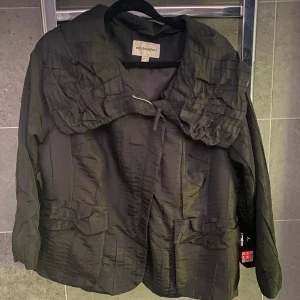 Svart jacka med volangkrage från easy comfort - Säljer en svart jacka, mörkare svart k verkligheten,  från easy comfort i storlek XL. Jackan har en unik volangkrage, dekorativa detaljer framtill och stängs med en knapp. Materialet har en lätt vågig snygg struktur och jackan har långa ärmar samt fickor framtill. 