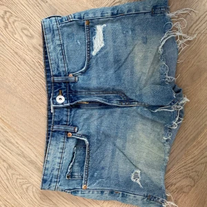 Blå jeansshorts med fransar - Säljer ett par blå jeansshorts med snyggt fransade kanter och slitningar både fram och bak. Klassisk femficksmodell med knappgylf och ljus tvätt från hm. Perfekta för sommaren! 