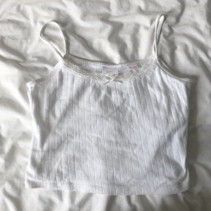Vitt ribbat linne - Säljer en söt vit ribbad linnetopp från Gina Tricot med tunna axelband och liten rosett framtill. Toppen har diskret mönster och spetsdetalj vid halsringningen. Perfekt för varma dagar eller som lager under andra plagg.