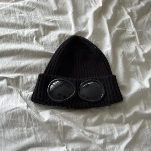 C.P. Company mössa  - Svart ribbstickad mössa från C.P. Company med inbyggda goggles framtill. Klassisk modell med uppvikt kant och tydlig logga på glasögonen. Perfekt accessoar för en cool och urban stil.